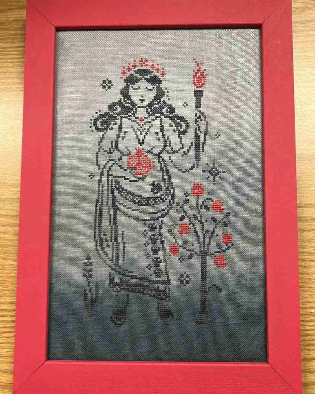 A framed cross-stitch artwork featuring a stylized female figure in a red crown and robe holding a torch, with red floral motifs on a gray background, set in a red frame on a wooden surface.