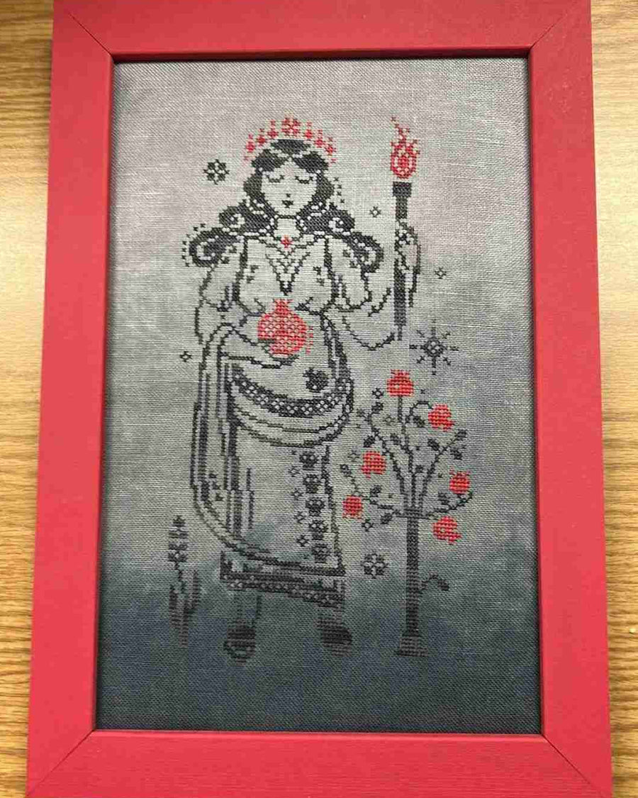 A framed cross-stitch artwork featuring a stylized female figure in a red crown and robe holding a torch, with red floral motifs on a gray background, set in a red frame on a wooden surface.
