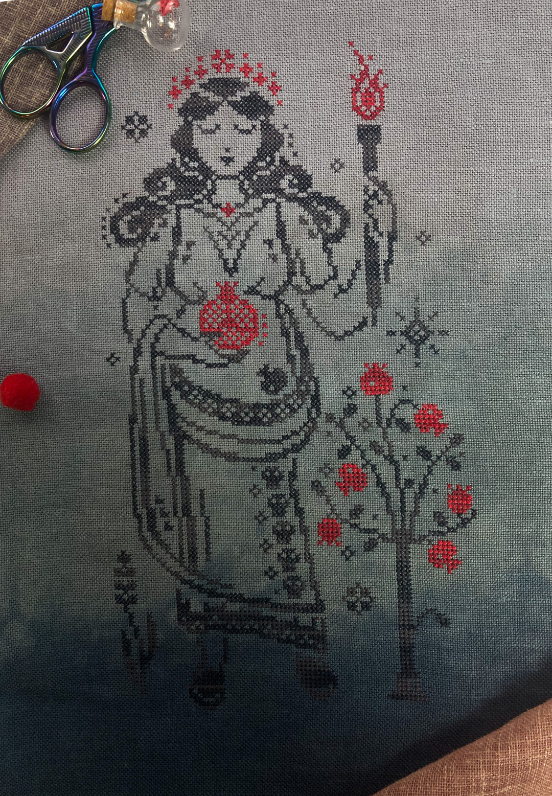 Cross-stitched design of a figure with red accents on gray fabric, scissors visible above.