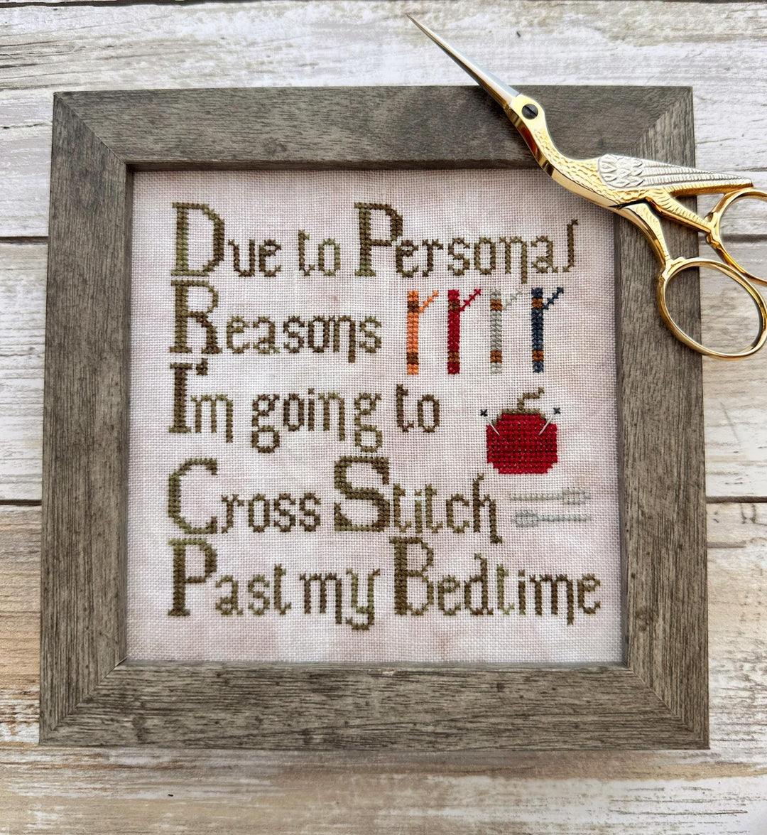 A framed cross-stitch artwork on a rustic wood surface, with a gold birdcage-style scissors prop resting on the frame. The stitched text reads, 'Due to Personal Reasons I'm going to Cross Stitch Past my Bedtime,' with small embroidered illustrations of a sewing machine, knitting needles, and a red apple.