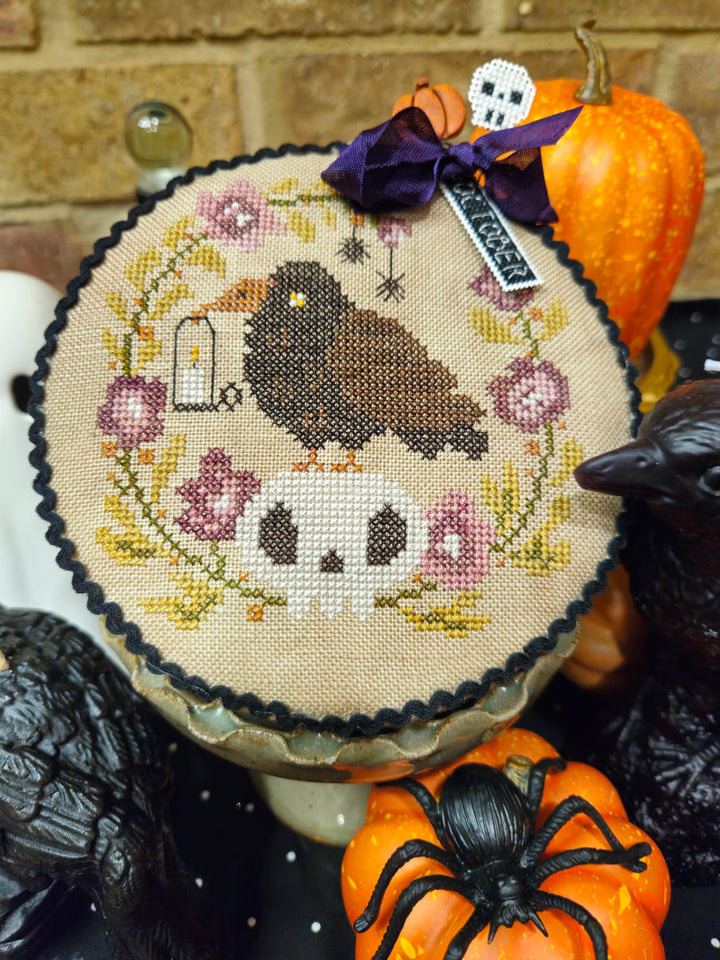 A round cross-stitch sampler featuring a raven, skull, and floral wreath design, styled with autumn props including pumpkins, a spider decoration, and a purple ribbon bow.