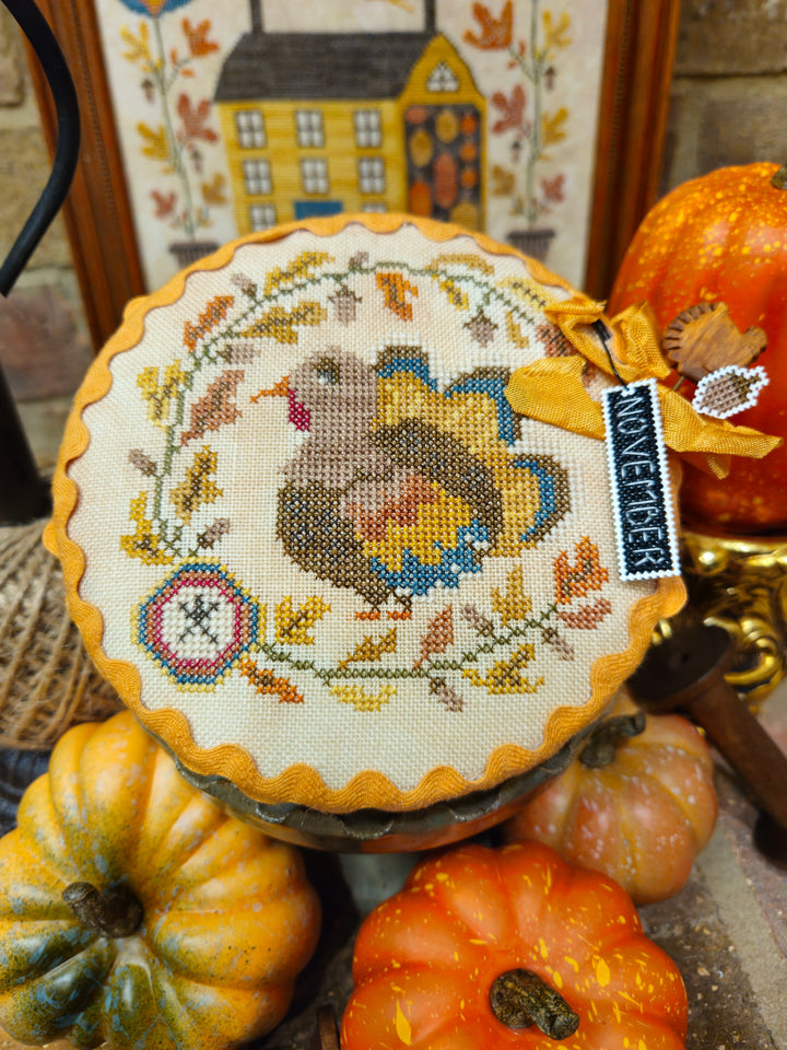 A round cross-stitch sampler featuring a turkey surrounded by autumn leaves and a house motif, displayed on a stand with decorative pumpkins and a small tag reading 'Novembers'.