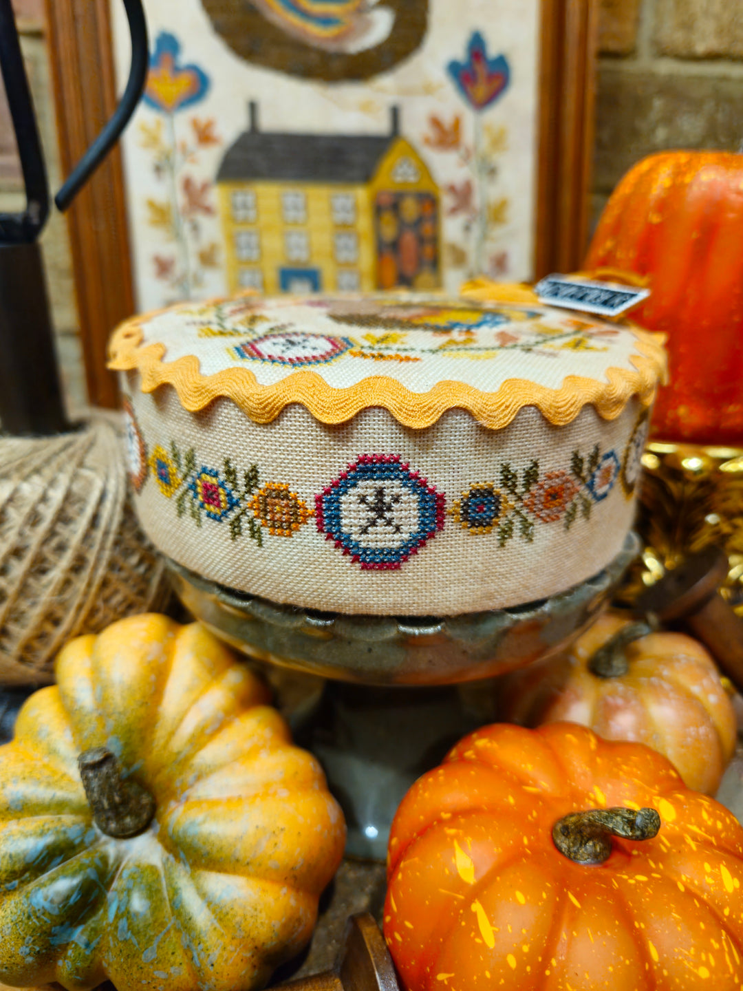 A round embroidered cushion or pouf with a scalloped yellow-orange trim and floral cross-stitch pattern, displayed on a stand among decorative pumpkins and autumn-themed decor.