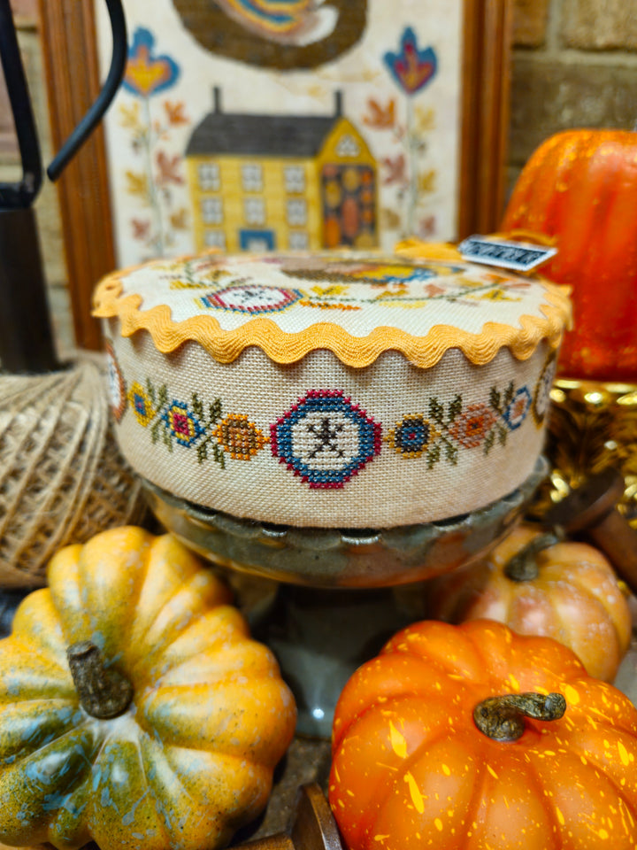 A round embroidered cushion or pouf with a scalloped yellow-orange trim and floral cross-stitch pattern, displayed on a stand among decorative pumpkins and autumn-themed decor.