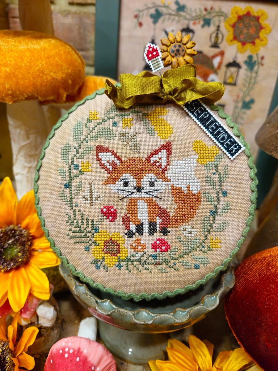 A round cross-stitch sampler featuring a fox surrounded by flowers and leaves, tied with a green ribbon and a tag that reads 'SPELENDER'. The sampler is displayed on a decorative stand among autumn-themed props including sunflowers and mushrooms.
