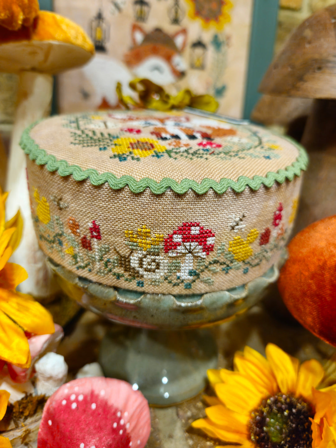 A round embroidered needlepoint canvas with a scalloped green edge, featuring a woodland mushroom and fox design, displayed on a glass stand among autumn-themed decorative props.