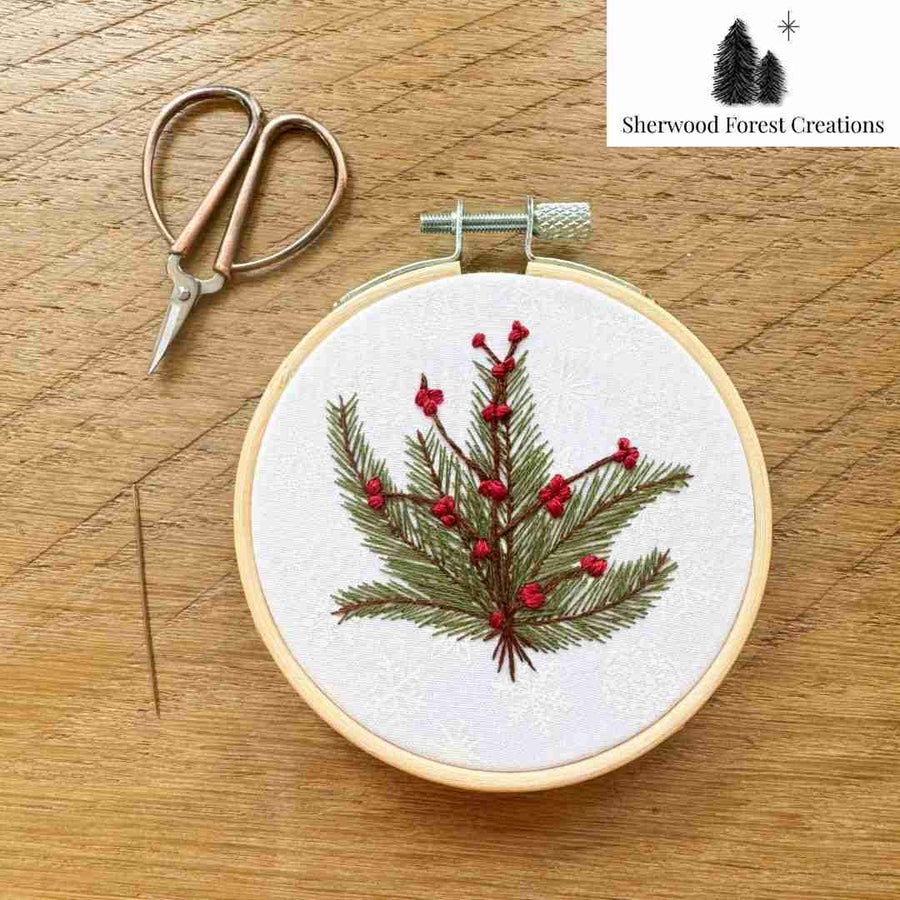 An embroidery hoop containing a green pine branch with red berries stitched on white fabric, placed on a wooden surface next to small metal embroidery scissors. A Sherwood Forest Creations logo appears in the top-right corner.