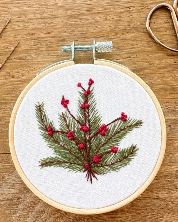 A close-up of a finished embroidery piece in a wooden hoop, featuring a green pine branch with red berries on a white fabric background, placed on a wooden tabletop with scissors partially visible.