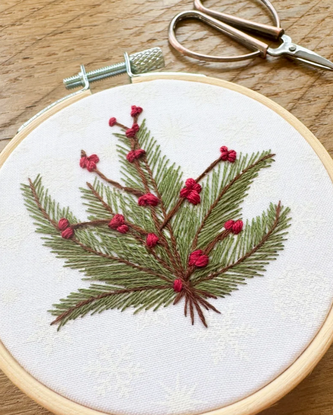 A close-up of a wooden embroidery hoop holding white fabric with a hand-embroidered pine branch design in olive green and red berries. A metal screw clamp is visible at the top, along with a pair of metal embroidery scissors on a wooden surface.