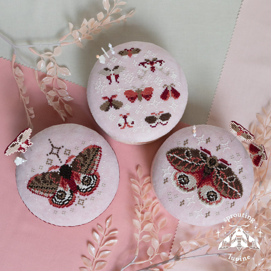 Three round cross-stitch sampler discs featuring embroidered moths and butterflies, arranged on a pink and pale green background with decorative dried leaves. A 'sprouting lupine' brand logo is visible in the bottom right corner.