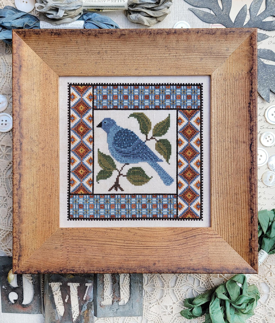 A framed cross-stitch artwork featuring a blue bird on a branch with green leaves, surrounded by geometric patterned borders. The piece is housed in a rustic wooden frame and styled on a textured fabric surface with decorative buttons and leaves.