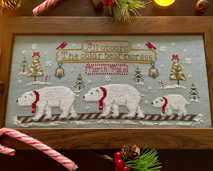 A framed Christmas cross-stitch piece featuring three polar bears wearing red scarves on a snowy path, with a banner reading 'All aboard The polar bear express' and 'North Pole'. The wooden frame is displayed on a dark wooden surface decorated with holiday elements like candy canes, pine branches, and a candle.