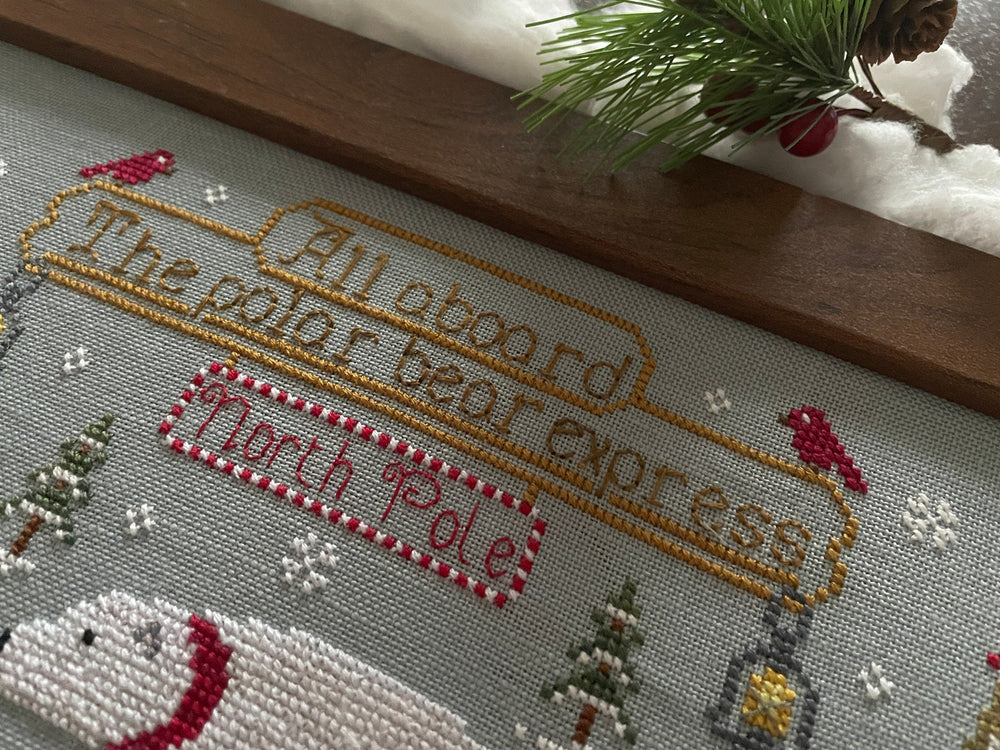 A close-up of a finished cross-stitch needlework piece featuring a polar bear, snowy trees, birds, and the embroidered text 'All aboard The polar bear express' and 'North Pole'.