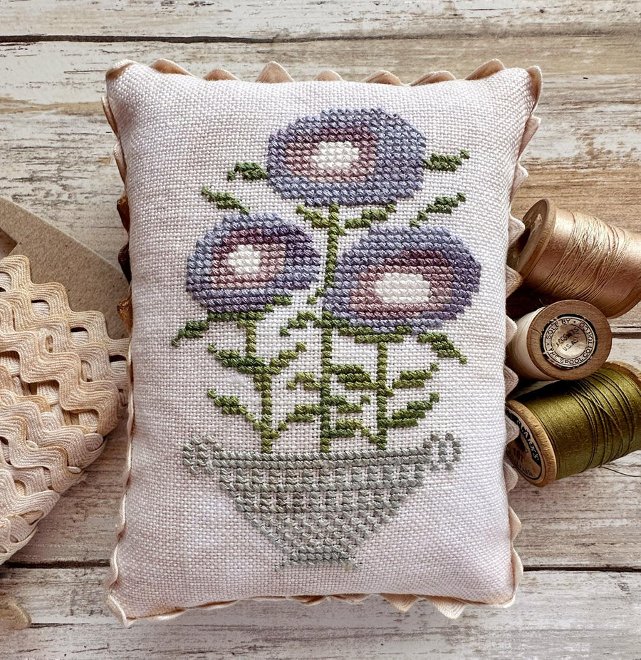 A square cross-stitch cushion with a floral design, styled on a rustic wooden surface with spools of thread and a wooden spoon.