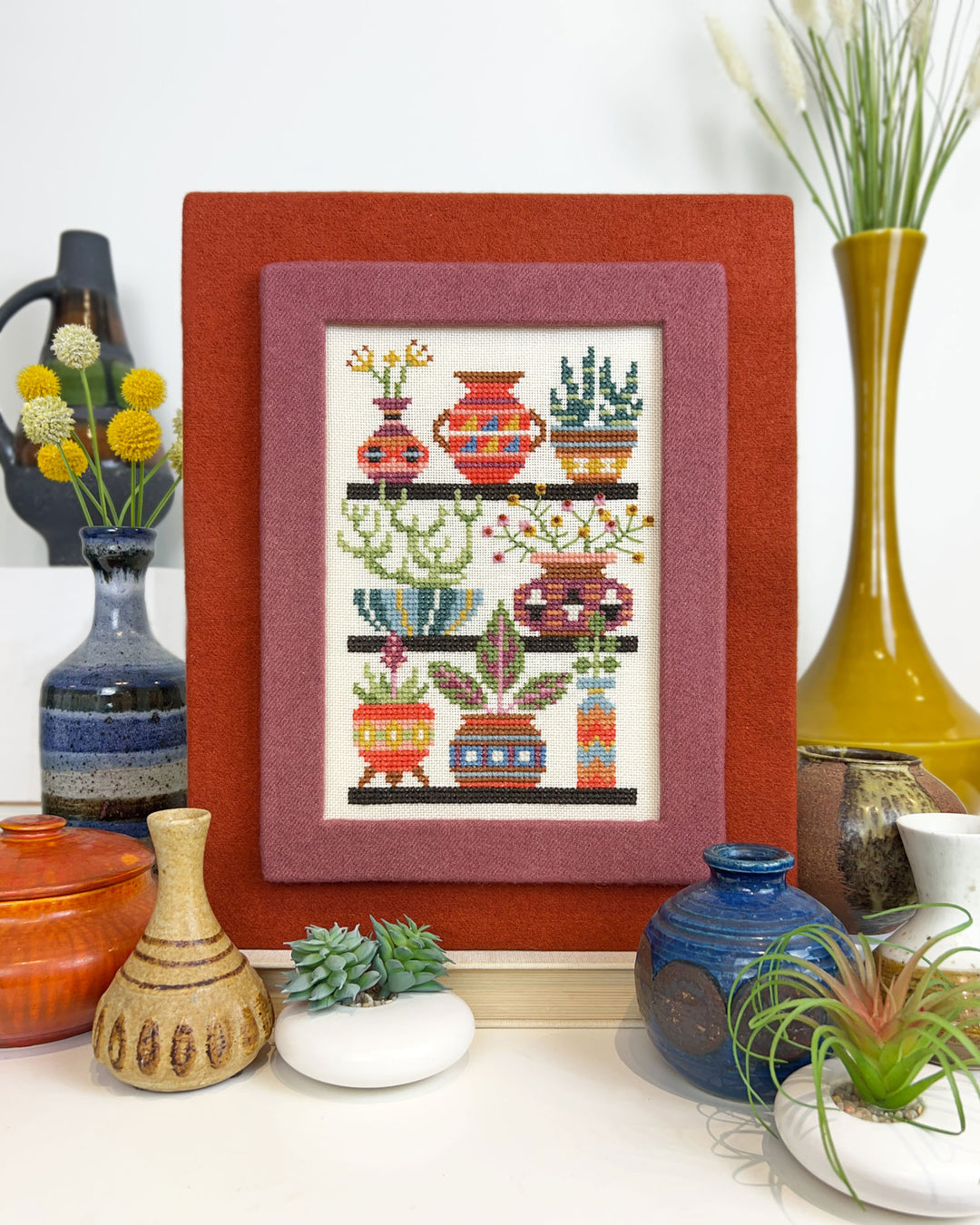 Framed cross-stitch art of colorful pots and plants on a shelf with decorative vases and plants.