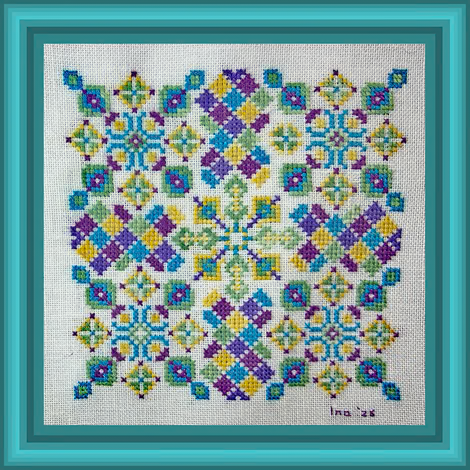 A framed square cross-stitch pattern featuring a colorful geometric floral design on white linen, surrounded by a teal wooden frame.