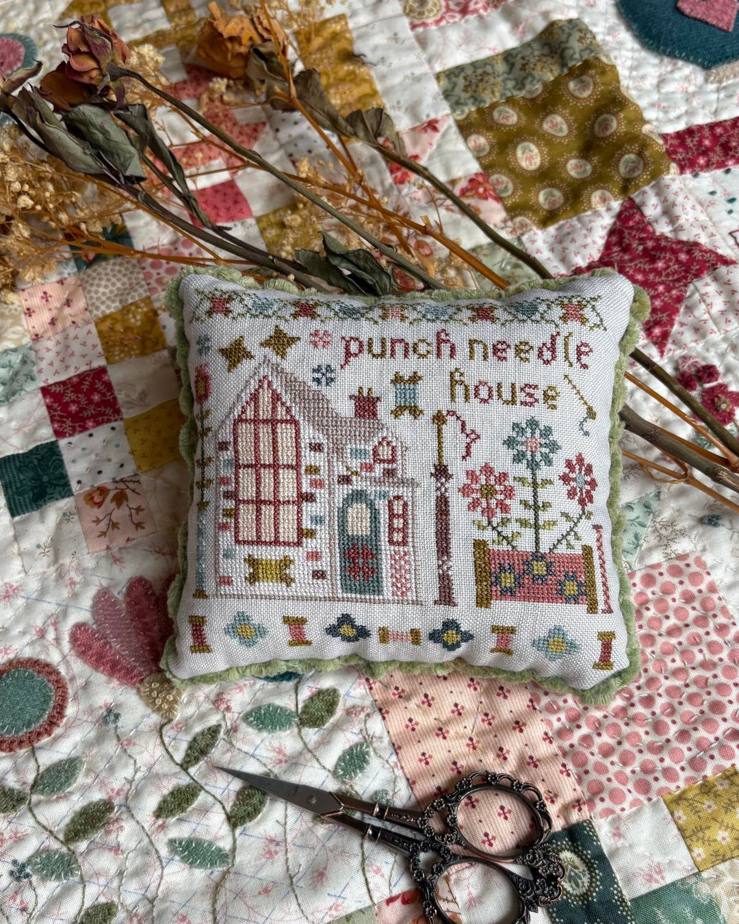 A small embroidered cross-stitch pillow with a house design and the words 'punch needle house' sits on a patchwork quilt, with dried flowers and embroidery scissors around it.