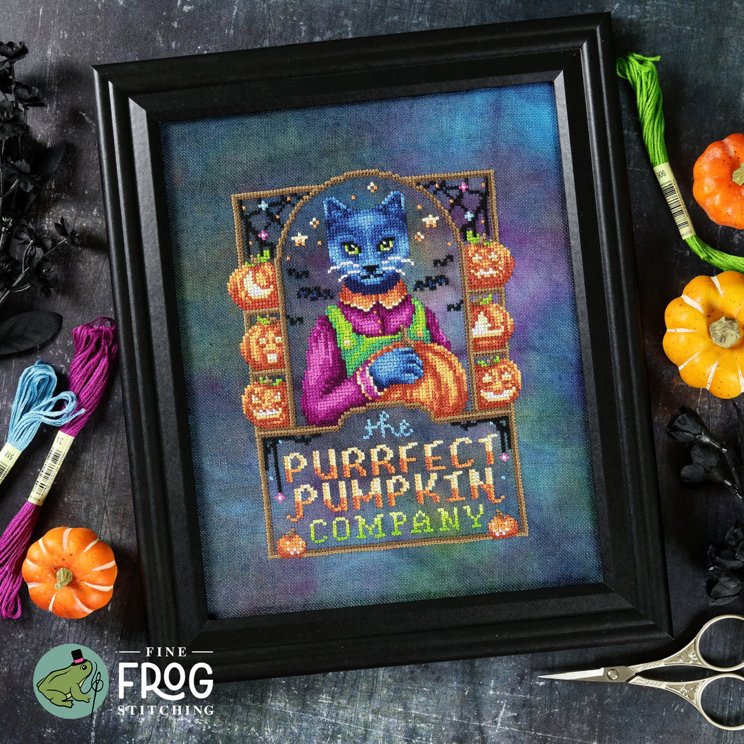 Framed cross-stitched picture of a cat with pumpkins and text 'The Purrfect Pumpkin Company' on a dark surface with embroidery threads and scissors.