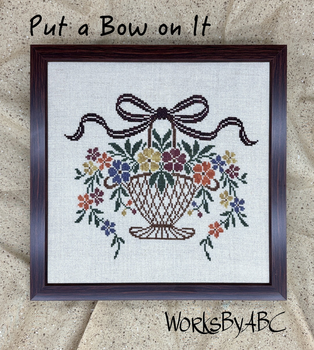 A framed cross-stitch design on linen fabric showing a basket of colorful flowers with a dark brown bow above it. Handwritten text at the top reads 'Put a Bow on It' and 'WorksByABC' appears at the bottom.