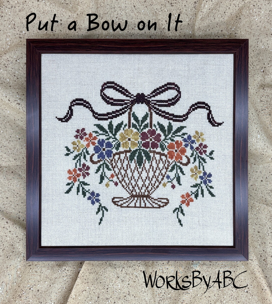 A framed cross-stitch design on linen fabric showing a basket of colorful flowers with a dark brown bow above it. Handwritten text at the top reads 'Put a Bow on It' and 'WorksByABC' appears at the bottom.