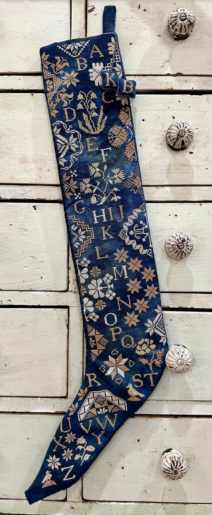 A navy blue Christmas stocking with gold embroidered letters, flowers, and botanical motifs, hanging against a rustic white wooden background with decorative knobs.