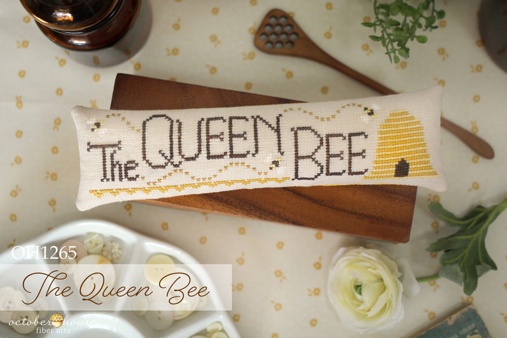 Cross-stitched pillow with 'The Queen Bee' design on a table with flowers and a bottle.
