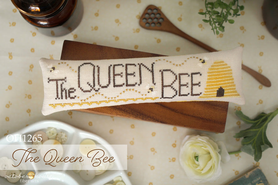 Cross-stitched pillow with 'The Queen Bee' design on a table with flowers and a bottle.
