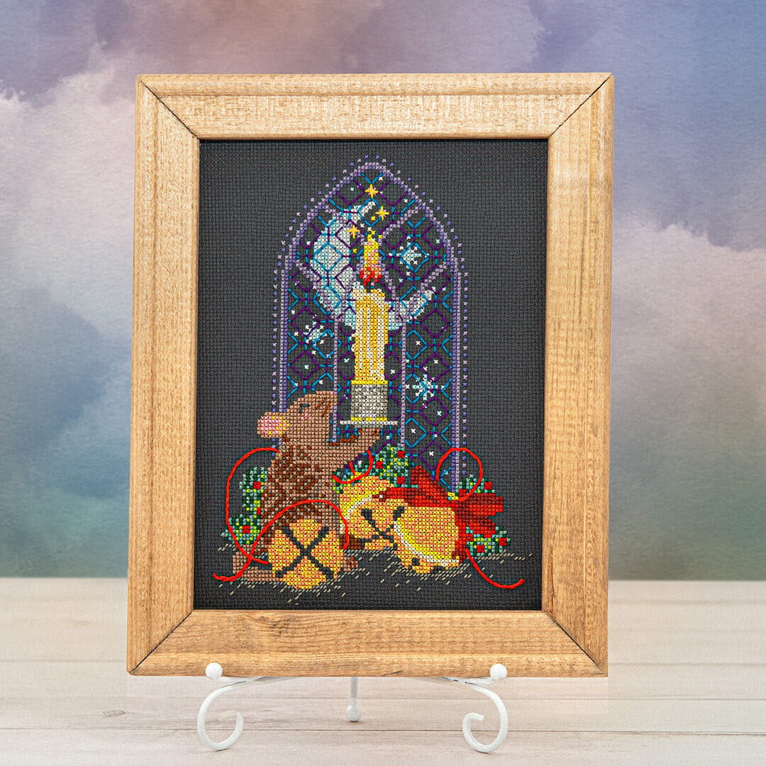 A framed needlepoint canvas featuring a Christmas scene with a mouse, a candle, and a gift box, displayed on a small white stand against a soft, cloudy sky background.
