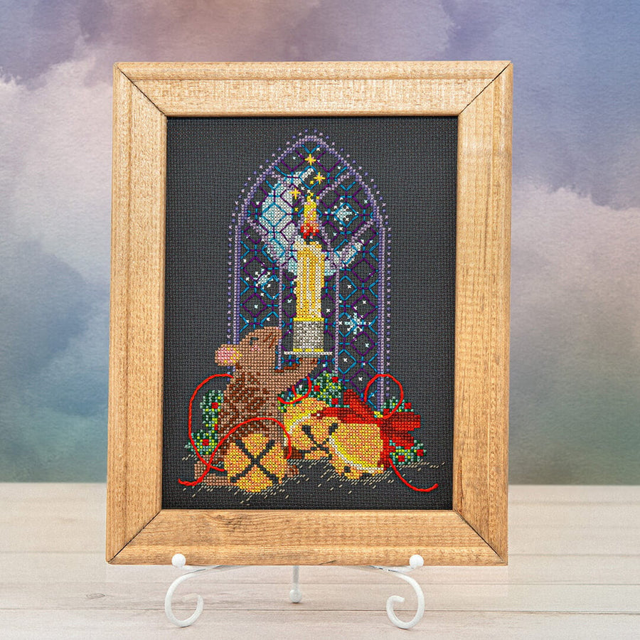 A framed needlepoint canvas featuring a Christmas scene with a mouse, a candle, and a gift box, displayed on a small white stand against a soft, cloudy sky background.