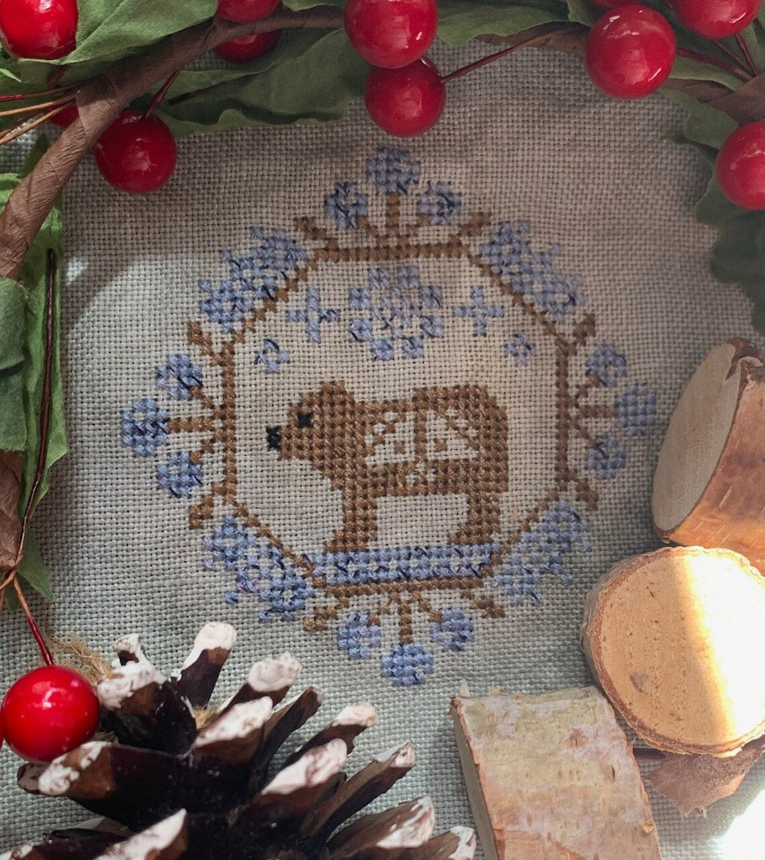 Cross-stitched design of a bear surrounded by berries and pinecones on fabric.