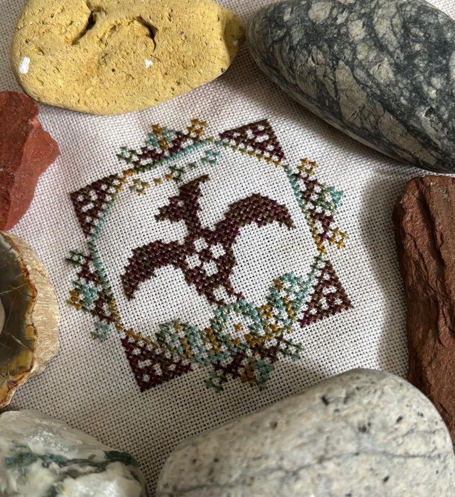 Cross-stitched design of a horse surrounded by decorative elements on fabric with rocks around.