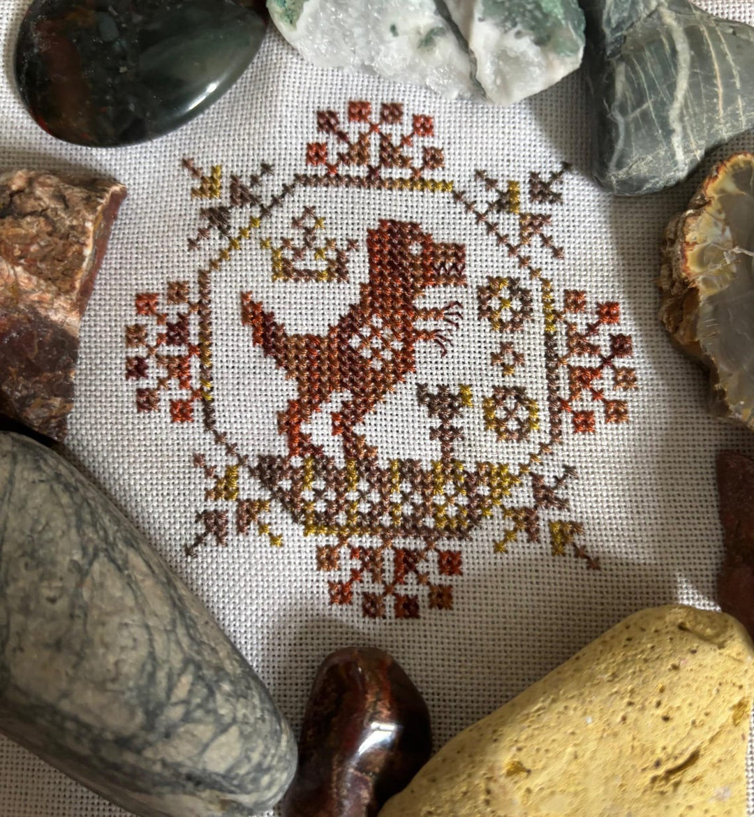 Cross-stitched design of a dog surrounded by decorative elements on fabric, surrounded by stones.