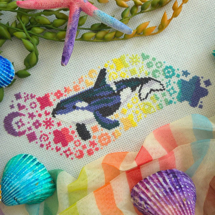 A close-up of a cross-stitch design featuring an orca whale surrounded by a rainbow of floral and wave patterns, styled with seashells, a starfish, and green seaweed on a white fabric background.
