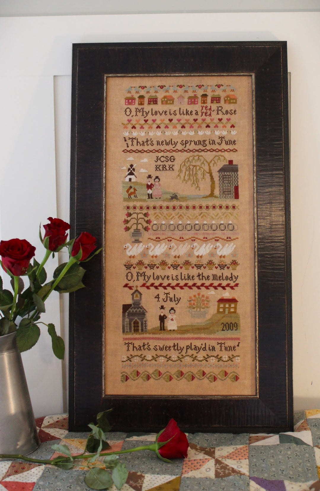 Framed cross-stitched sampler with decorative elements on a quilted surface.