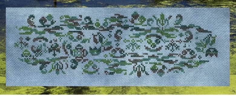 A rectangular needlepoint canvas featuring a horizontal band of stylized green and brown botanical or tribal-like motifs on a light blue-gray mesh background, shown against a blurred yellow and dark natural backdrop.