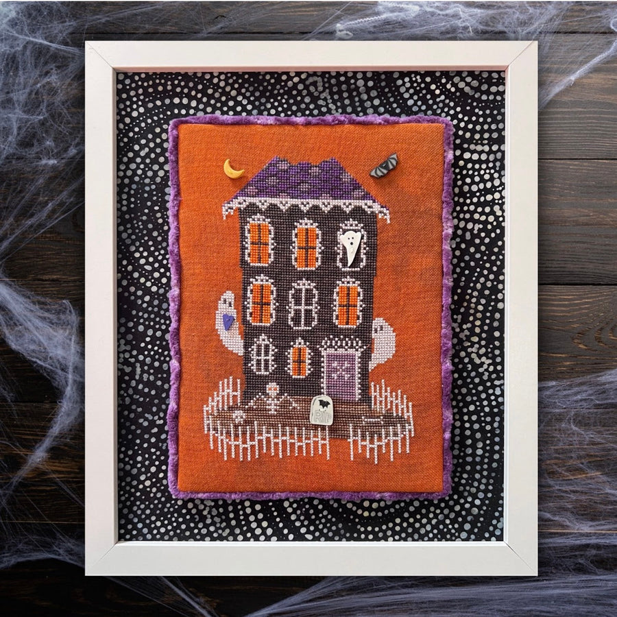 Framed cross-stitched Halloween house on an orange background with spider webs.
