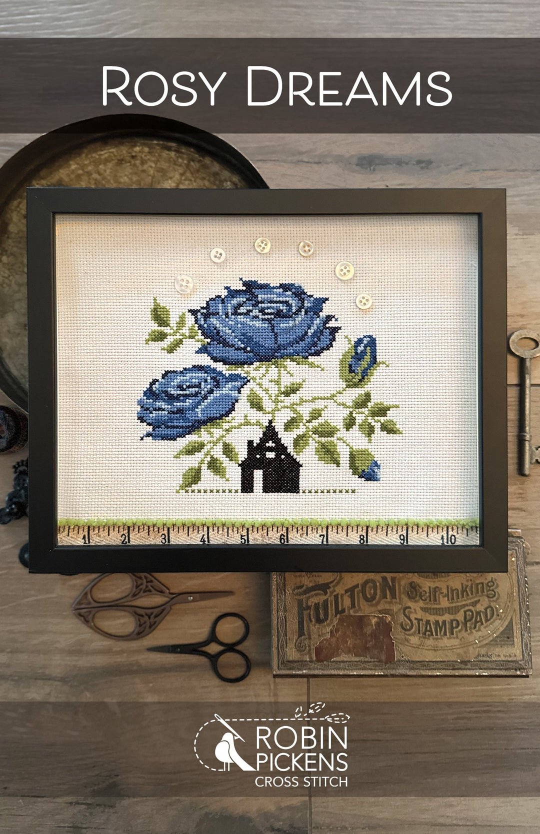 Cross-stitched design of blue roses and a house on fabric, framed and displayed with sewing tools.