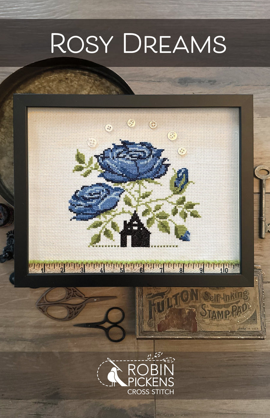 Cross-stitched design of blue roses and a house on fabric, framed and displayed with sewing tools.