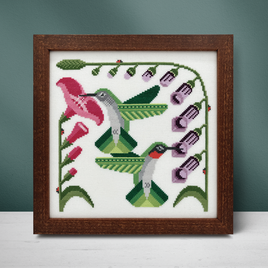 Framed cross-stitched artwork of hummingbirds and flowers on a white background