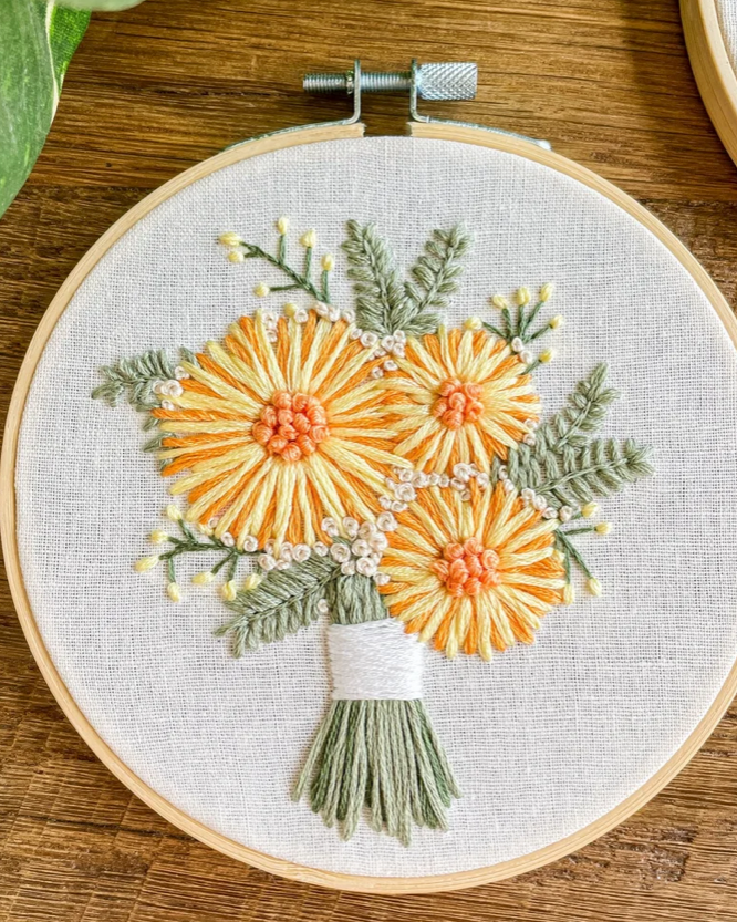 A close-up of a wooden embroidery hoop holding a floral bouquet design stitched on textured off-white fabric. The bouquet features three large yellow-orange flowers with peach centers, green leaves, and a white wrapped stem, photographed on a wooden surface.