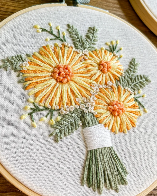 A close-up of a hand-embroidered floral bouquet in a wooden hoop embroidery sampler. The bouquet features three yellow and orange flowers with green leaves and stems, tied with white thread, on a beige linen fabric background.