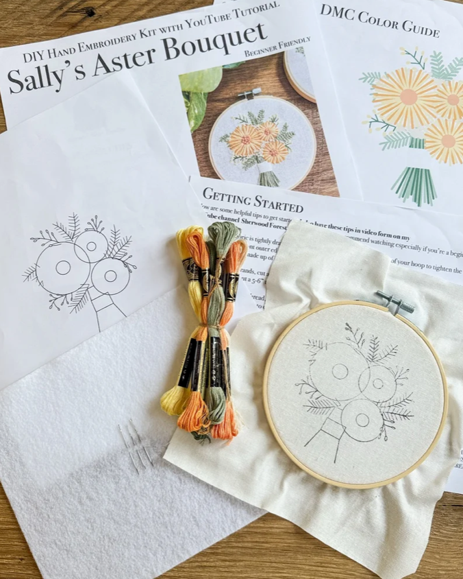 Embroidery kit components arranged on a wooden surface, including printed pattern sheets titled 'Sally's Aster Bouquet' and 'DMC Color Guide,' a bundle of multicolor embroidery floss, a white fabric piece with a floral outline, and an embroidery hoop with matching fabric.