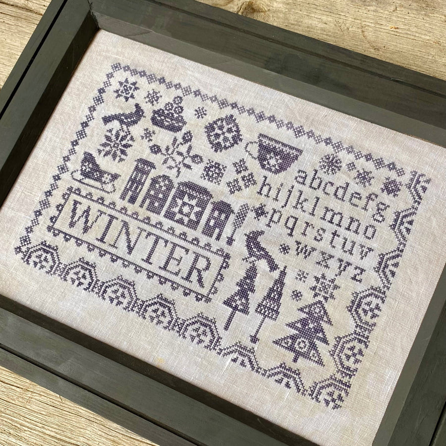 Framed cross-stitched pattern with winter-themed designs on a wooden surface