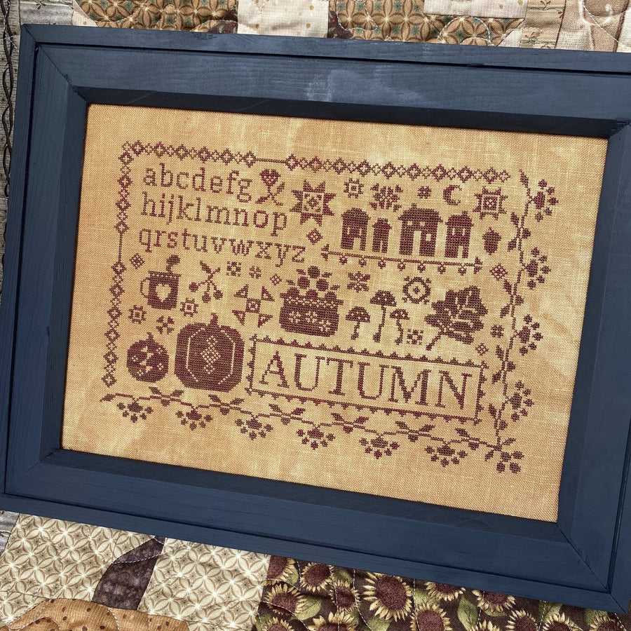 Framed cross-stitched sampler with 'Autumn' text and decorative elements on a textured fabric background.