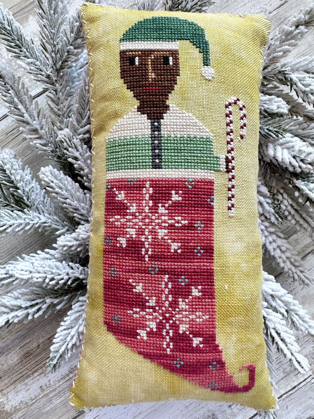 Decorative pillow with a cross-stitched design of an elf in a green hat and red stocking with snowflake pattern, surrounded by flocked branches.