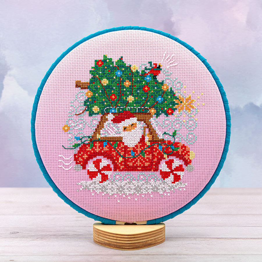 A circular cross-stitch embroidery hoop on a wooden stand, featuring a festive design of a Santa Claus in a red car carrying a decorated Christmas tree, set against a soft pastel sky background.