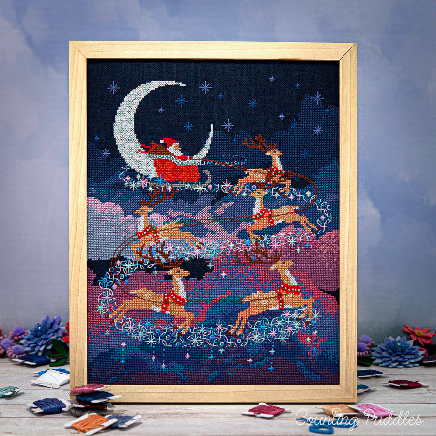 Framed cross-stitched artwork of Santa Claus in a sleigh with reindeer against a night sky.