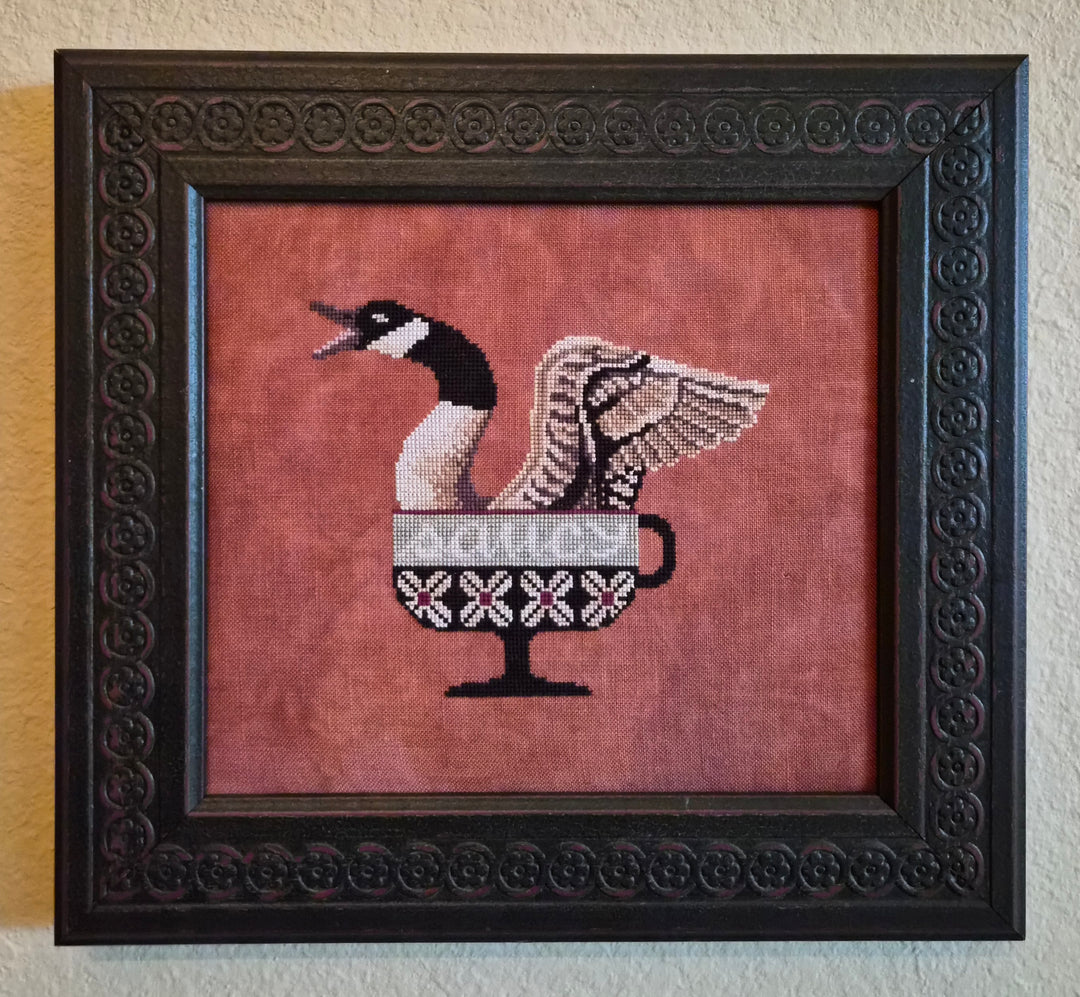 Framed cross-stitched artwork of a goose with a cup on a red background