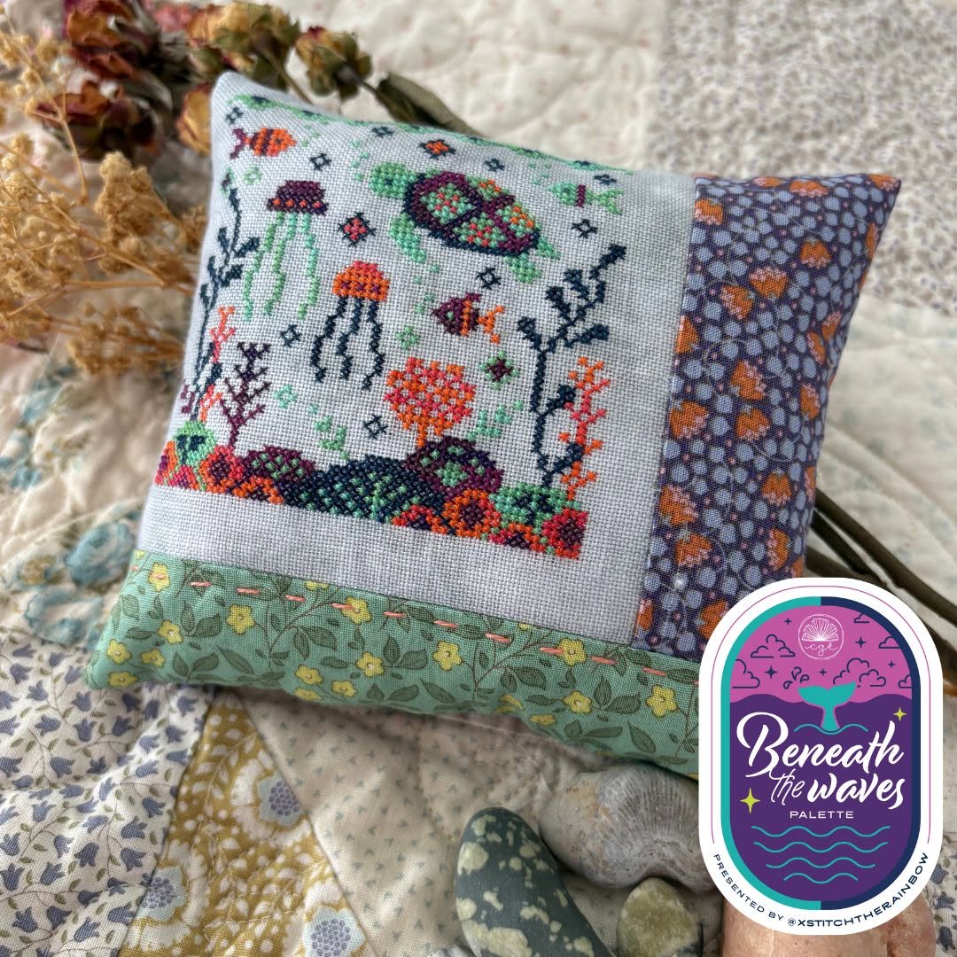Cross-stitched pillow with ocean-themed design on a textured surface, featuring the 'Beneath the Waves' palette label.