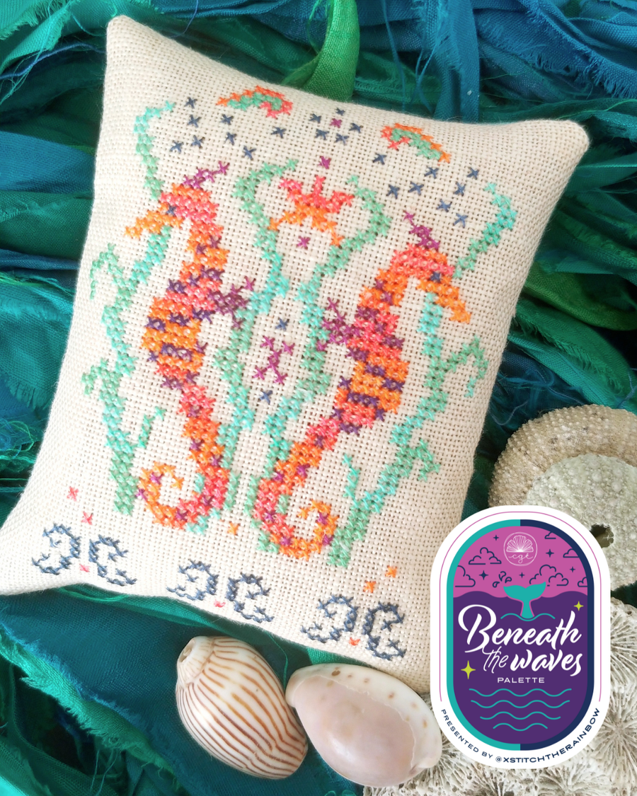 Cross-stitched pillow with seahorses on a textured green fabric background, featuring the 'Beneath the Waves Palette' logo.
